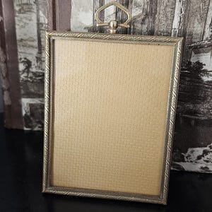 May include: A rectangular gold-toned metal picture frame featuring a textured border with a repeating diagonal pattern. The frame includes a decorative pointed arch-shaped hanging loop at the top and contains a solid gold-colored fabric insert.