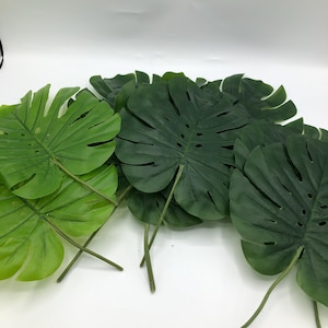 May include: A collection of artificial monstera leaves featuring deep green and lighter green shades. Each leaf has a distinct split-leaf pattern with visible veins and stems, arranged in a overlapping cluster to display the foliage texture.