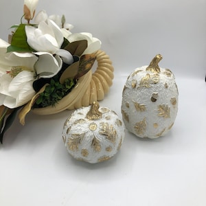 May include: Two white pumpkins with textured surfaces and metallic gold leaf accents shaped like oak leaves and acorns, accompanied by a cream-colored cornucopia overflowing with white magnolia flowers and green foliage.