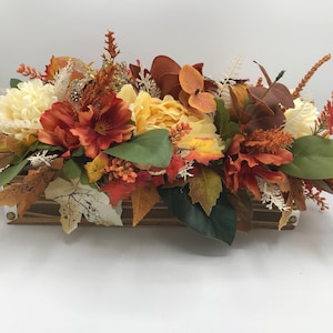 May include: A wooden box centerpiece with fall-themed floral arrangements. The arrangement features white, yellow, and orange flowers, along with green and brown leaves.