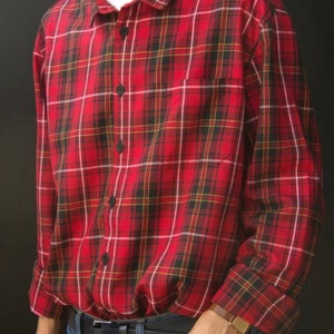 May include: Red plaid button-down shirt with black buttons and a chest pocket. The shirt has a classic plaid pattern in red, black, yellow, and white. The shirt is worn with blue jeans and a black belt.