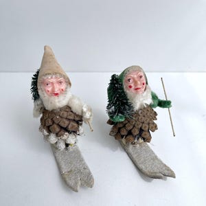 Op de afbeelding: Twee vintage kerstkabouterornamenten. Elke kabouter heeft een kegelvormig lichaam, een geschilderd gezicht en een hoed. Eén kabouter draagt een beige hoed, de andere een groene hoed. Ze zijn bevestigd aan grijze ski's en hebben kleine bomen.