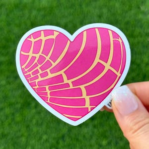 May include: A heart-shaped sticker featuring a pink concha design with yellow accents. The sticker has a white border and is held against a green grassy background.
