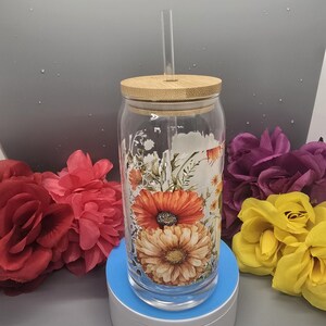 Puede incluir: Un vaso de vidrio transparente con un dise&ntilde;o floral con flores naranjas, amarillas y blancas. El vaso tiene una tapa de bamb&uacute; y una pajita de pl&aacute;stico transparente. El vaso est&aacute; sobre una base de carga azul y blanca.