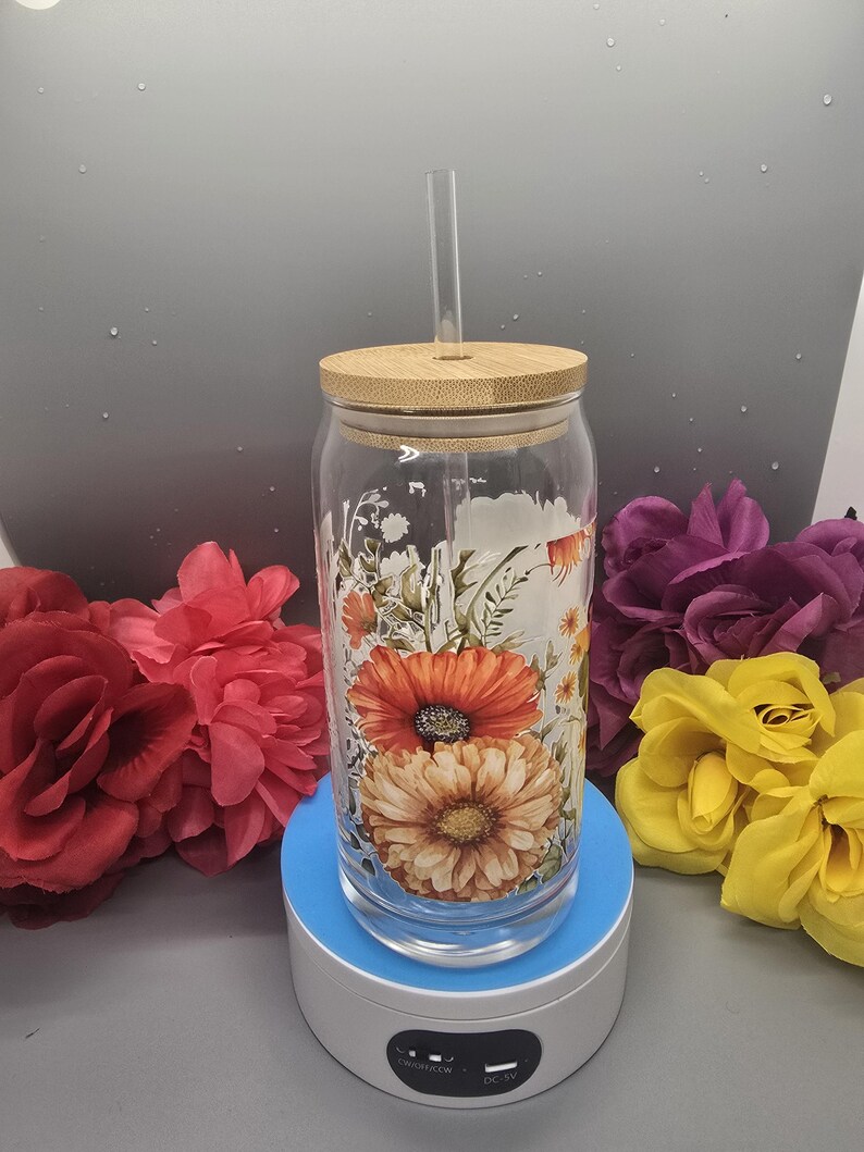Puede incluir: Un vaso de vidrio transparente con un dise&ntilde;o floral con flores naranjas, amarillas y blancas. El vaso tiene una tapa de bamb&uacute; y una pajita de pl&aacute;stico transparente. El vaso est&aacute; sobre una base de carga azul y blanca.