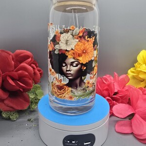 Puede incluir: Un vaso de vidrio con un dise&ntilde;o floral que presenta a una mujer con una corona de flores. El vaso tiene una tapa de bamb&uacute; y una pajita. El vaso est&aacute; en un soporte de exhibici&oacute;n giratorio con una base azul y una parte superior blanca. El soporte tiene un interruptor con el texto "CW/OFF/CCW" y un puerto USB con el texto "DC-5V".