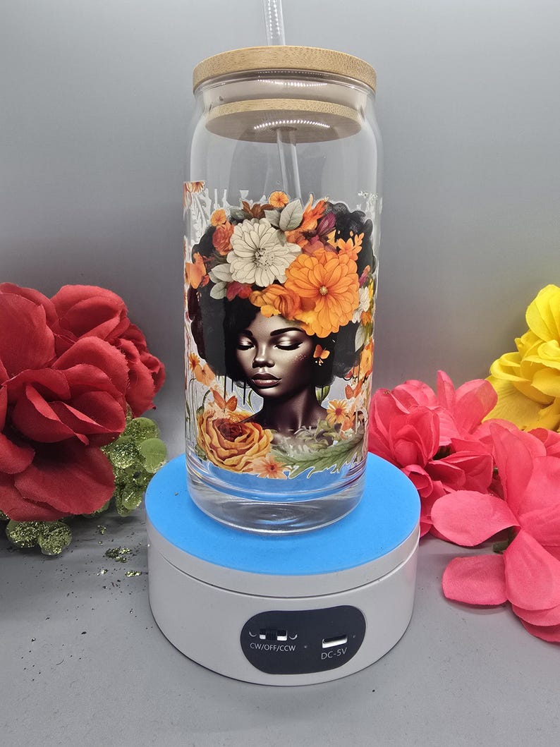 Puede incluir: Un vaso de vidrio con un dise&ntilde;o floral que presenta a una mujer con una corona de flores. El vaso tiene una tapa de bamb&uacute; y una pajita. El vaso est&aacute; en un soporte de exhibici&oacute;n giratorio con una base azul y una parte superior blanca. El soporte tiene un interruptor con el texto "CW/OFF/CCW" y un puerto USB con el texto "DC-5V".
