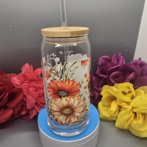 Puede incluir: Un vaso de vidrio transparente con tapa de bamb&uacute; y una pajita. El vaso tiene un dise&ntilde;o floral con flores naranjas, amarillas y blancas. El vaso est&aacute; sobre una base giratoria blanca.