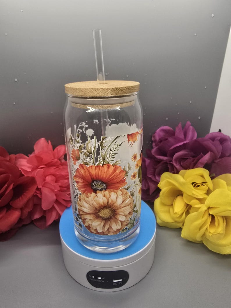 Puede incluir: Un vaso de vidrio transparente con tapa de bamb&uacute; y una pajita. El vaso tiene un dise&ntilde;o floral con flores naranjas, amarillas y blancas. El vaso est&aacute; sobre una base giratoria blanca.