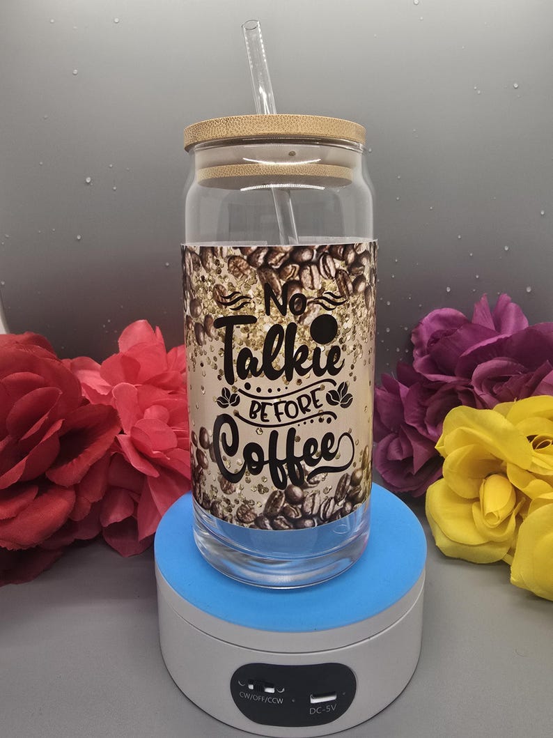 Puede incluir: Un vaso de vidrio transparente con tapa de bamb&uacute; y una pajita. El vaso tiene un fondo de purpurina dorada con un gr&aacute;fico negro que dice "No Talkie Before Coffee".