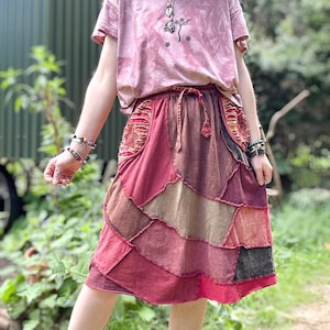 Op de afbeelding: Een roze tie-dye t-shirt met een klein bloemenontwerp en een patchwork rok in tinten rood, bruin en bruin. De rok heeft een tailleband met trekkoord en een gelaagd, asymmetrisch ontwerp. De outfit wordt buiten gedragen.