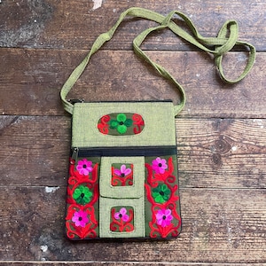Puede incluir: Bolso bandolera verde oliva con diseños florales bordados en rosa, rojo y verde. El bolso tiene una correa larga y ajustable y un compartimento principal con cremallera. Forma rectangular con bolsillo frontal.