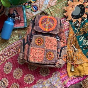 Puede incluir: Una mochila de patchwork marrón con un símbolo de paz bordado en la parte delantera. La mochila tiene detalles en naranja, rojo y marrón. La mochila está sobre una tela estampada de colores.