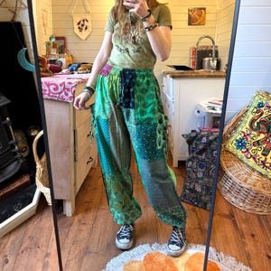 Puede incluir: Pantalones verdes de patchwork con una variedad de patrones y texturas. Los pantalones tienen cintura con cordón y bolsillos. La persona lleva una camiseta verde y zapatillas negras. El estilo general es bohemio.