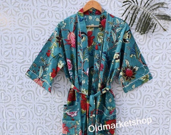 Puede incluir: Una bata de algodón azul turquesa con estampado floral y ribetes blancos. La bata tiene un cinturón de atar y dos bolsillos. La bata está colgada de una percha de madera sobre un fondo blanco con un patrón floral.