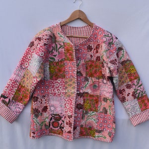 May include: A pink quilted jacket with a floral patchwork design. The jacket has a V-neck and a button closure. The sleeves are long and have a striped cuff. The text "old market shop" is visible on the jacket.