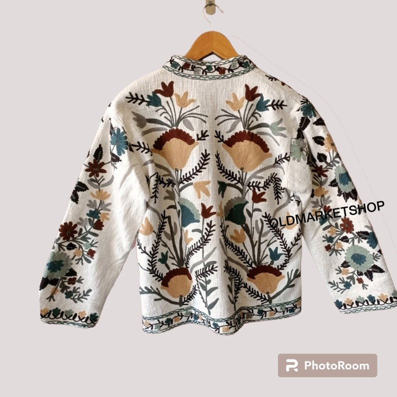 Puede incluir: Chaqueta blanca bordada con motivos florales en tonos marrones, verdes y beige. La chaqueta tiene un cuello mandar&iacute;n y mangas largas. El texto "OLDMARKETSHOP" es visible en la manga derecha.