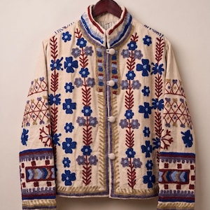 May include: A cream-coloured jacket featuring a high collar and four round buttons. The garment is decorated with intricate embroidery in shades of blue, red, and gold, displaying floral patterns, vertical stems, and geometric border designs.