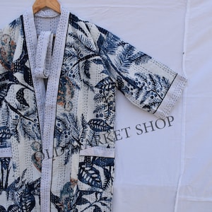 May include: A white kimono-style robe with a blue botanical print featuring leaves and palm trees. The robe has a white trim and is hanging on a wooden hanger. The text "OLD MARKET SHOP" is visible on the right side of the robe.