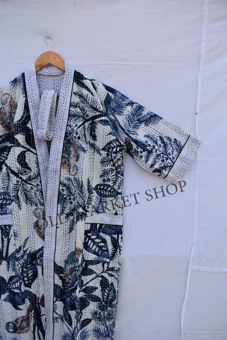 May include: A white kimono-style robe with a blue botanical print featuring leaves and palm trees. The robe has a white trim and is hanging on a wooden hanger. The text "OLD MARKET SHOP" is visible on the right side of the robe.