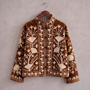 May include: A brown velvet jacket featuring cream-colored floral embroidery throughout the front and sleeves. The jacket has a high collar, long sleeves, and a front closure secured by four round brown fabric-covered buttons on a vertical placket.