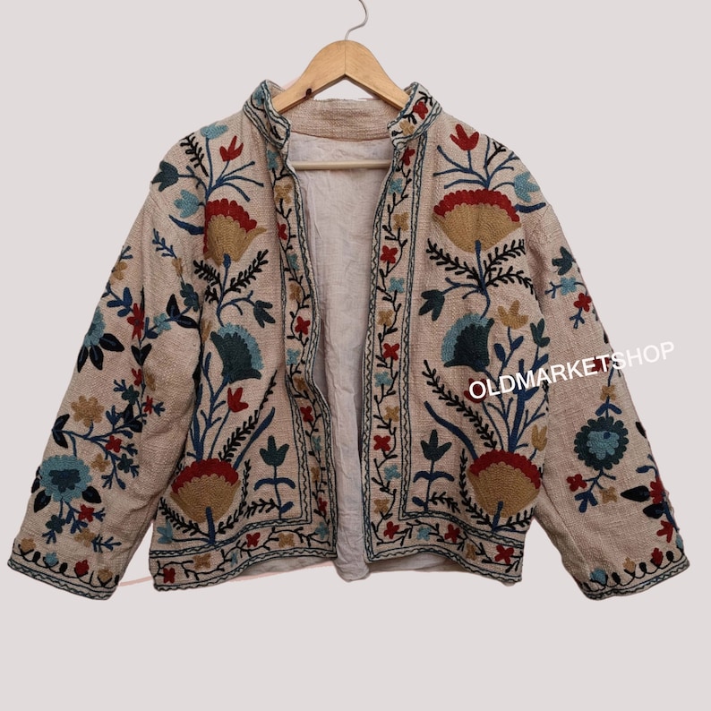 Puede incluir: Una chaqueta beige con un dise&ntilde;o de bordado floral. La chaqueta tiene un cuello mandar&iacute;n y mangas largas. El bordado es en tonos de azul, rojo, verde y amarillo.
