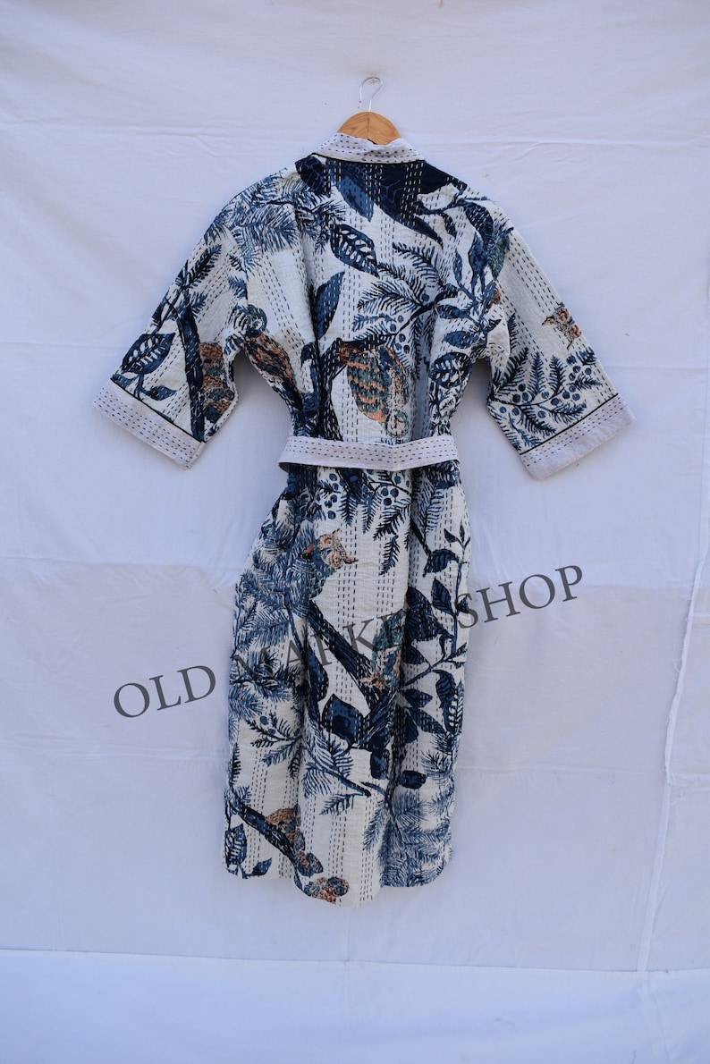 May include: White robe with a blue and tan botanical print. The robe features a matching belt and short sleeves. The fabric is patterned with leaves, birds, and other natural elements. The robe is hanging on a wooden hanger.