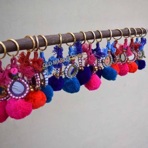 May include: A collection of colorful keychains with pom-poms and small mirrors. Each keychain features a different color combination of pink, red, blue, and white. The keychains are hanging on a wooden rod.
