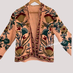 Puede incluir: Una chaqueta rosa bordada con un patr&oacute;n floral en tonos de verde, azul, rojo y marr&oacute;n. La chaqueta tiene manga larga y cuello en V. El texto "OLDMARKETSHOP" es visible en el lado derecho de la chaqueta.