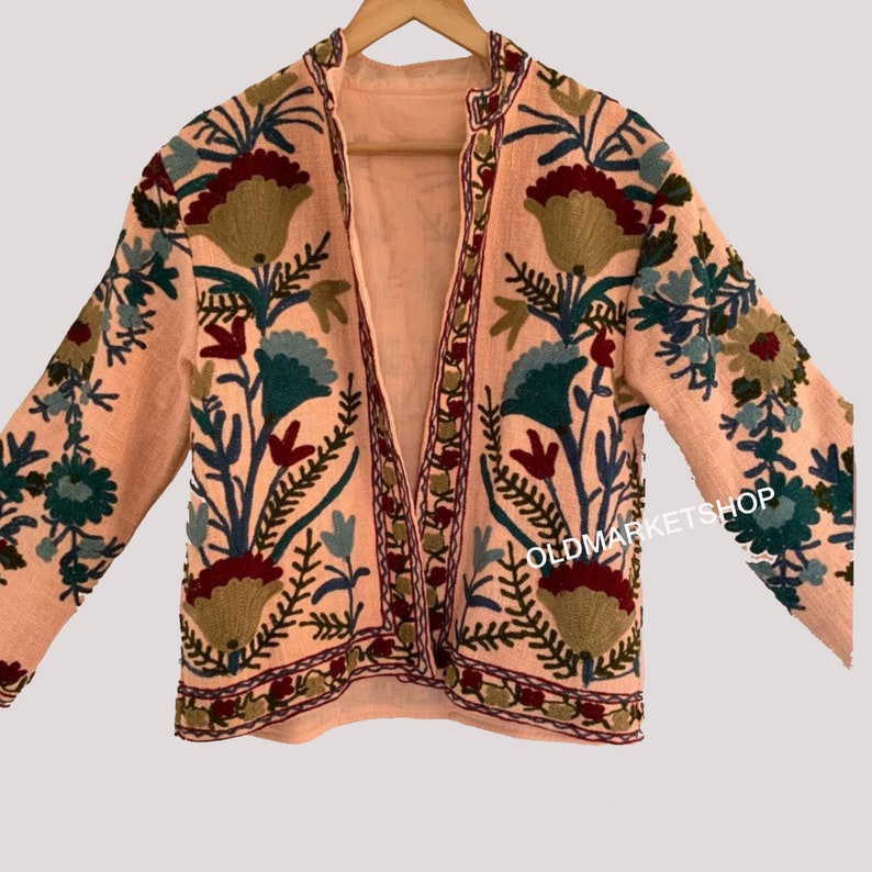 Puede incluir: Una chaqueta rosa bordada con un patr&oacute;n floral en tonos de verde, azul, rojo y marr&oacute;n. La chaqueta tiene manga larga y cuello en V. El texto "OLDMARKETSHOP" es visible en el lado derecho de la chaqueta.