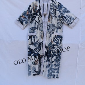 May include: A white robe with a blue and beige floral pattern, hanging on a wooden hanger. The robe has a shawl collar, pockets, and a matching belt. The text "OLD MARKET SHOP" is visible on the bottom.