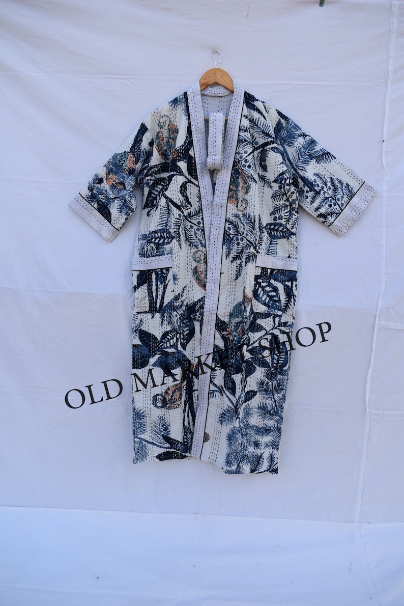 May include: A white robe with a blue and beige floral pattern, hanging on a wooden hanger. The robe has a shawl collar, pockets, and a matching belt. The text "OLD MARKET SHOP" is visible on the bottom.