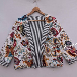 Pode incluir: Casaco acolchoado com padrão floral em tons de vermelho, azul, amarelo e laranja sobre fundo branco. O interior e a barra do casaco apresentam riscas a preto e branco. O casaco está pendurado num cabide de madeira.