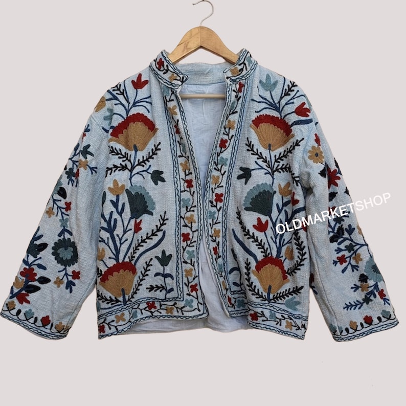 Puede incluir: Una chaqueta azul claro bordada con motivos florales en rojo, azul, verde y marr&oacute;n. La chaqueta tiene un cuello mandar&iacute;n y un cierre frontal.