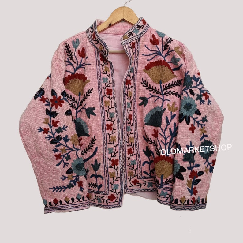 Puede incluir: Una chaqueta rosa bordada con motivos florales en tonos azules, rojos y marrones. La chaqueta tiene un cuello mandar&iacute;n y una cremallera frontal. El texto "OLDMARKETSHOP" es visible en la esquina inferior derecha de la chaqueta.