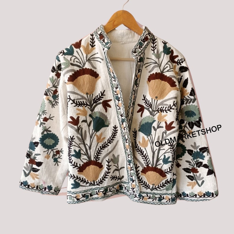 Puede incluir: Una chaqueta de lino blanca con un dise&ntilde;o de bordado floral en tonos marrones, verdes y beige. La chaqueta tiene un cuello mandar&iacute;n y un cierre de botones en la parte delantera. El texto "OLDMARKETSHOP" es visible en la esquina inferior derecha de la imagen.