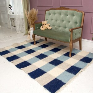 May include: A checkered rug in shades of blue, white, and beige is placed in front of a green velvet sofa with a teddy bear sitting on it. The sofa is in a room with white walls and a pink wall behind it.