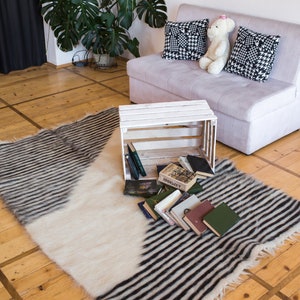 K&ouml;nnte beinhalten: Eine wei&szlig;e Holzkiste mit darin gestapelten B&uuml;chern steht auf einem schwarz-wei&szlig; gestreiften Teppich. Der Teppich liegt auf einem Holzboden. Im Hintergrund befindet sich eine wei&szlig;e Couch mit schwarz-wei&szlig; gemusterten Kissen.