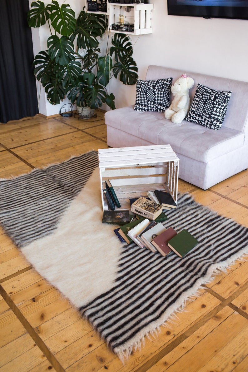 K&ouml;nnte beinhalten: Eine wei&szlig;e Holzkiste mit darin gestapelten B&uuml;chern steht auf einem schwarz-wei&szlig; gestreiften Teppich. Der Teppich liegt auf einem Holzboden. Im Hintergrund befindet sich eine wei&szlig;e Couch mit schwarz-wei&szlig; gemusterten Kissen.