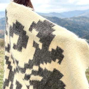 May include: A white and gray woven blanket with a geometric pattern. The blanket is draped over a person's shoulders, with the background showing a mountain range.