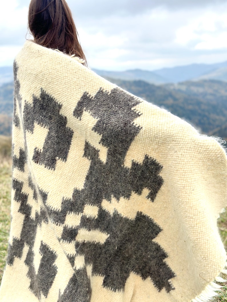 May include: A white and gray woven blanket with a geometric pattern. The blanket is draped over a person's shoulders, with the background showing a mountain range.