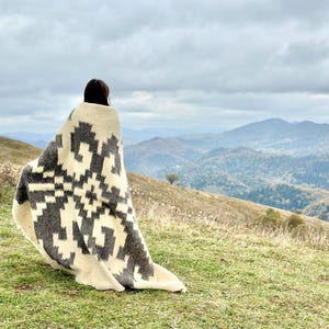 May include: A person wrapped in a white and black patterned blanket stands on a grassy hill overlooking a mountain range. The sky is cloudy and the mountains are covered in trees.