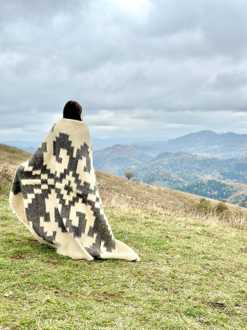 May include: A person wrapped in a white and black patterned blanket stands on a grassy hill overlooking a mountain range. The sky is cloudy and the mountains are covered in trees.