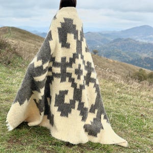 May include: A person wearing a white and gray patterned blanket with a geometric design. The blanket is made of wool and has a soft, cozy feel. The person is standing in a field with a mountain range in the background.