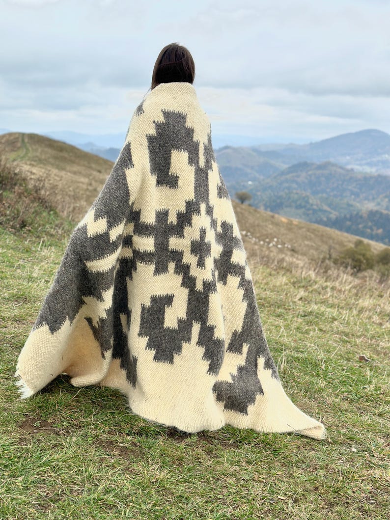 May include: A person wearing a white and gray patterned blanket with a geometric design. The blanket is made of wool and has a soft, cozy feel. The person is standing in a field with a mountain range in the background.