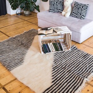 K&ouml;nnte beinhalten: Ein wei&szlig;-schwarz gestreifter Teppich mit flauschiger Textur. Der Teppich liegt auf einem Holzboden vor einem wei&szlig;en Sofa mit schwarz-wei&szlig;en gemusterten Kissen und einem Teddyb&auml;r.