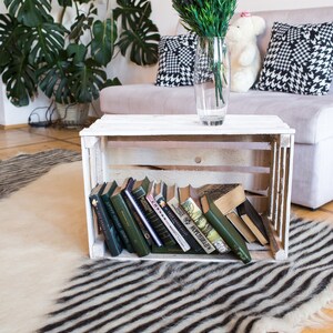 K&ouml;nnte beinhalten: Eine wei&szlig;e Holzkiste mit einer Auswahl an B&uuml;chern, die darin gestapelt sind. Die Kiste steht auf einem schwarz-wei&szlig; gestreiften Teppich. Im Hintergrund steht ein Tisch mit einer Vase voller lila Blumen.