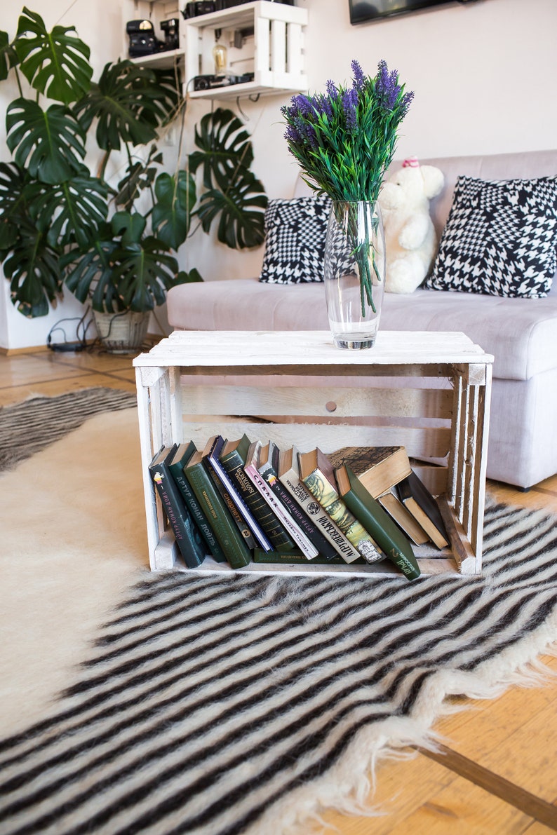 K&ouml;nnte beinhalten: Eine wei&szlig;e Holzkiste mit einer Auswahl an B&uuml;chern, die darin gestapelt sind. Die Kiste steht auf einem schwarz-wei&szlig; gestreiften Teppich. Im Hintergrund steht ein Tisch mit einer Vase voller lila Blumen.
