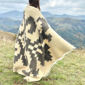 May include: A person wearing a white and gray patterned blanket with a fringe border. The blanket is draped over the person's shoulders and is flowing in the wind. The person is standing on a grassy hill with a mountain range in the background.