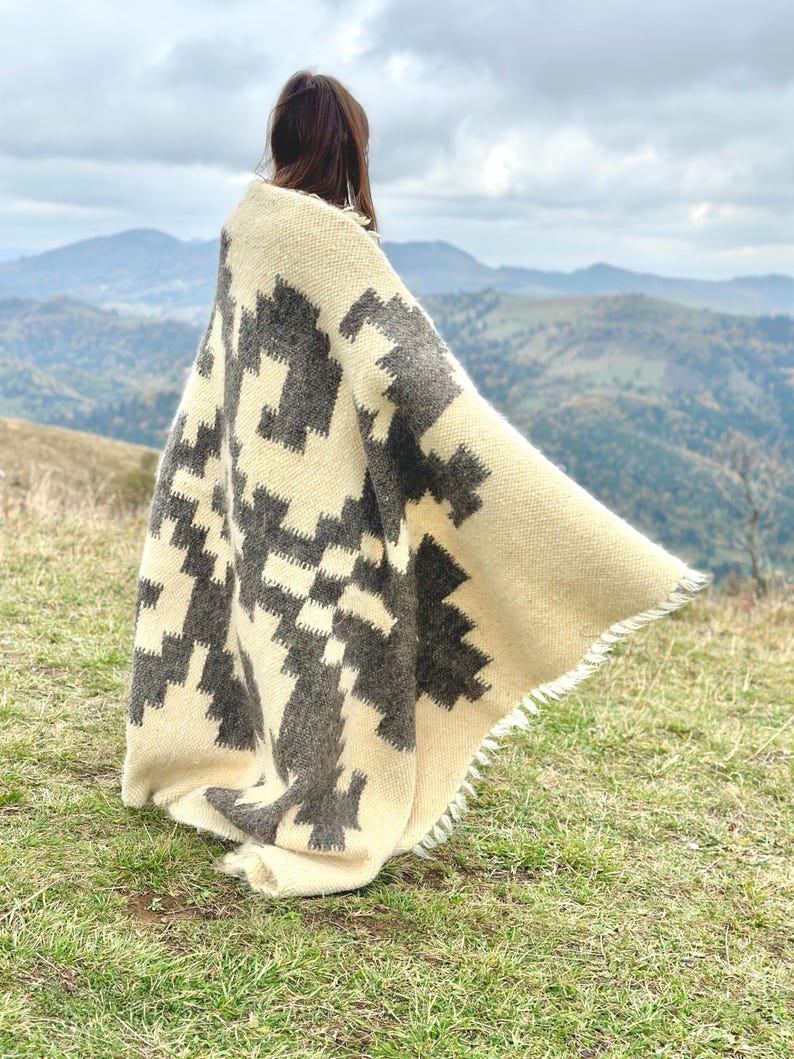 May include: A person wearing a white and gray patterned blanket with a fringe border. The blanket is draped over the person's shoulders and is flowing in the wind. The person is standing on a grassy hill with a mountain range in the background.